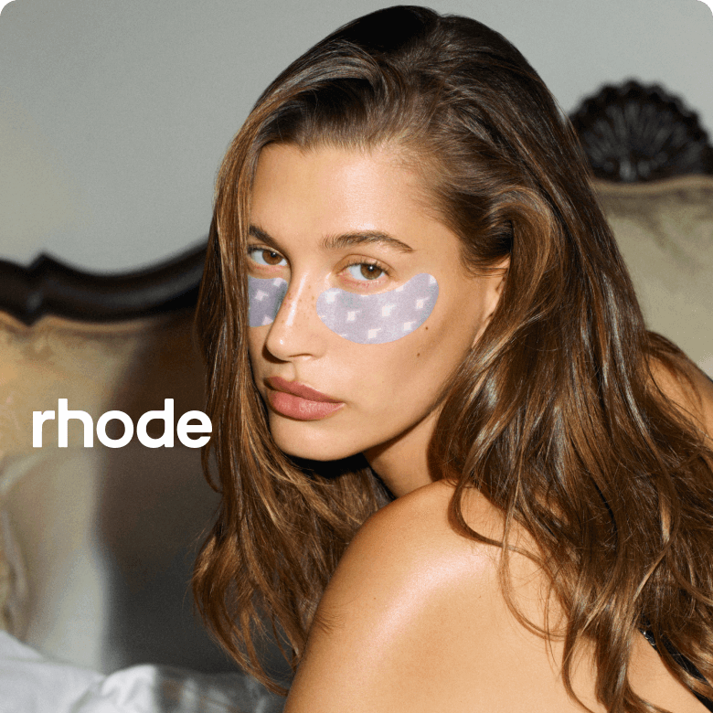 Woman with eye patches sitting on a bed, wearing a black top, with 'rhode' branding.