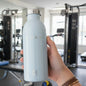 Hand holding a light blue water bottle with a brand logo in a gym setting