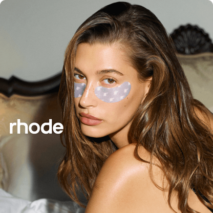 Woman with eye patches sitting on a bed, wearing a black top, with 'rhode' branding.