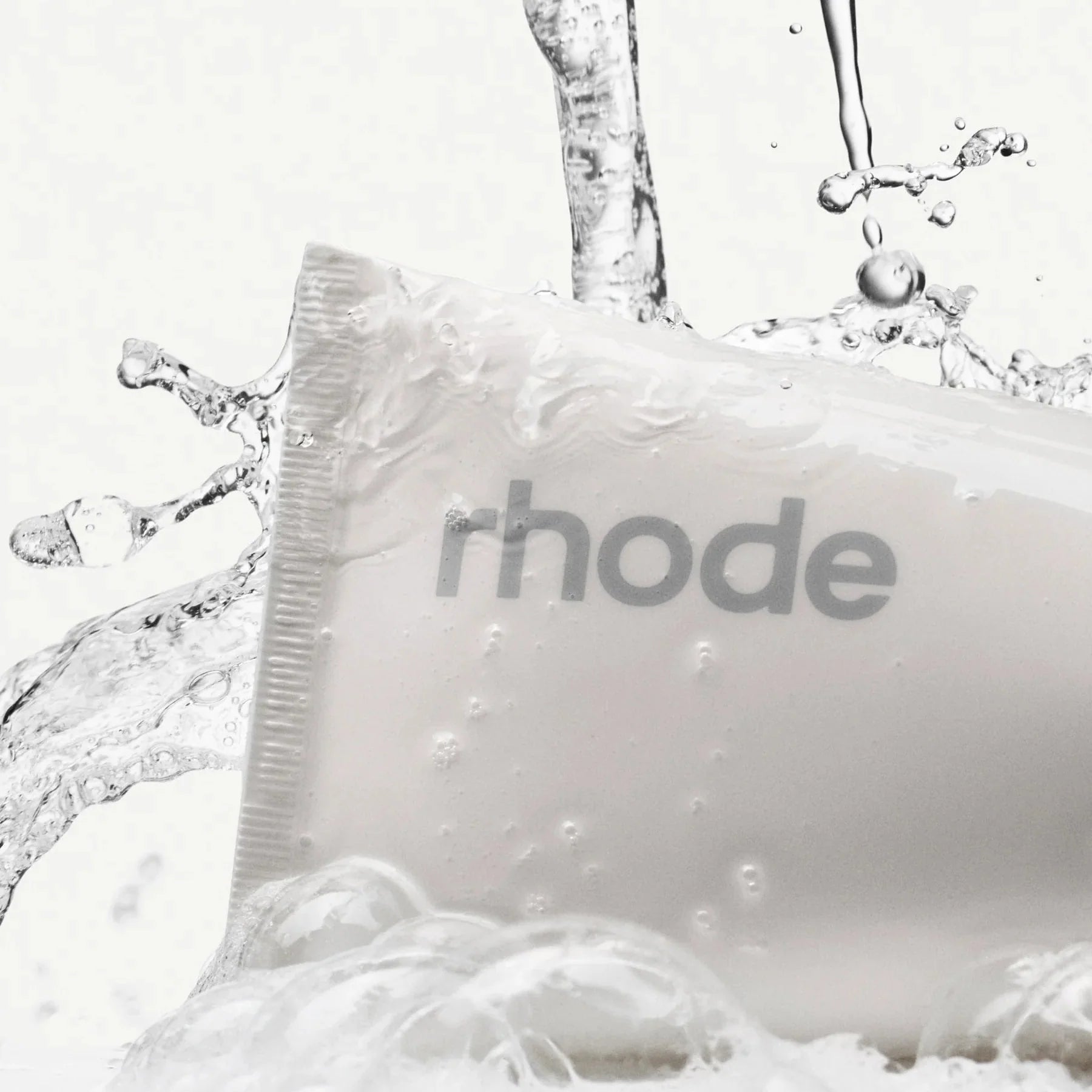White bar of soap with 'rhode' branding surrounded by water droplets on a white background